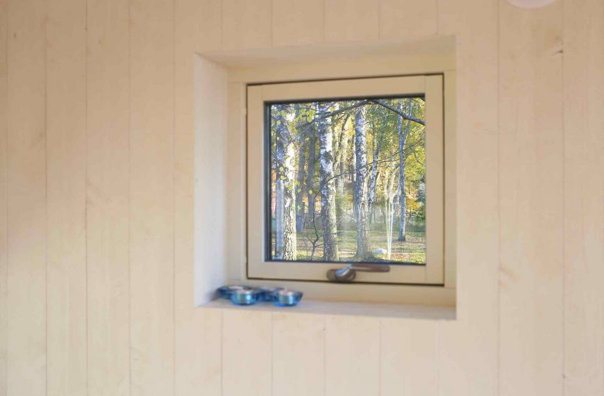 Litet fönster i en vitmålad trävägg. Utanför syns björkskogen. Attefallare formgiven av Anders Frelin och Lilla Sthlm.