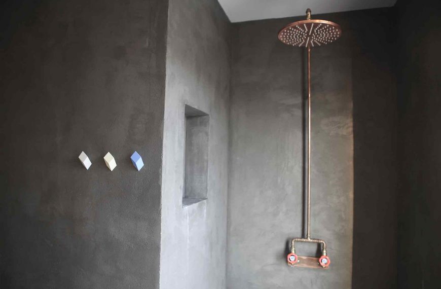 Badrum med väggar i betong. I duschnischen sitter en takdusch i koppar med röda vred. Arkitekt är Anders Frelin.