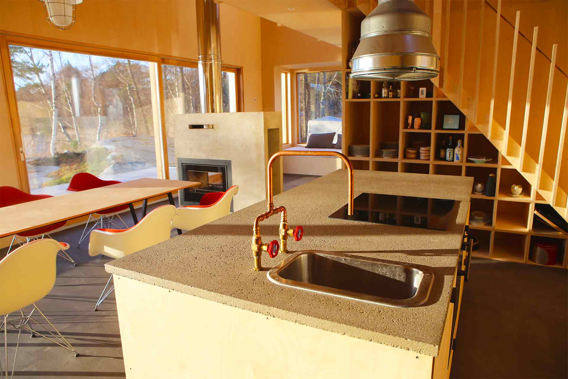 En köksö i björkplywood med betongbänk. Bakom syns fritidshusets stora skjutdörrar i glas. Utanför glaset syns vattnet.