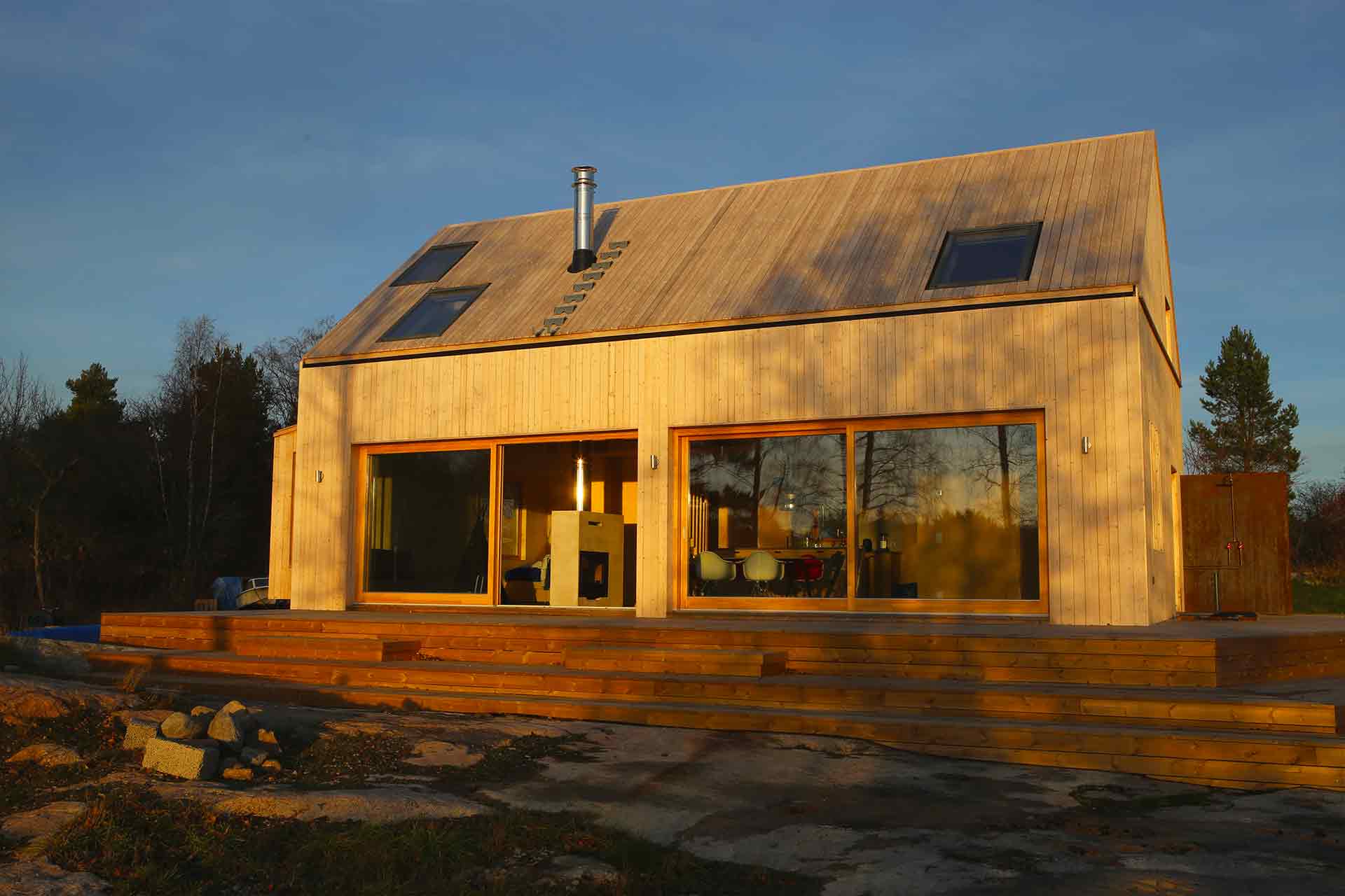 En modern lada med träpanel på både väggar och tak. I stora skjutfönster reflekteras skogen och vattnet framför huset.