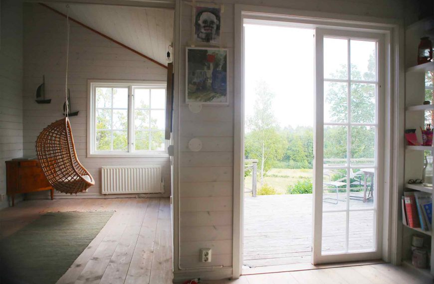 En hängande korgstol i ett rum med liggande vit panel. Genom fönstren syns äng och björkskog i skärgårdsmiljö.